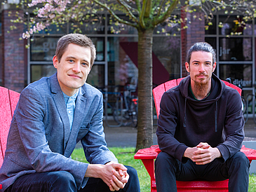 Two young men sit on red chairs in park and look at camera