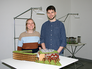 Young woman and young man standing in front of a table with a 3D model of a building.